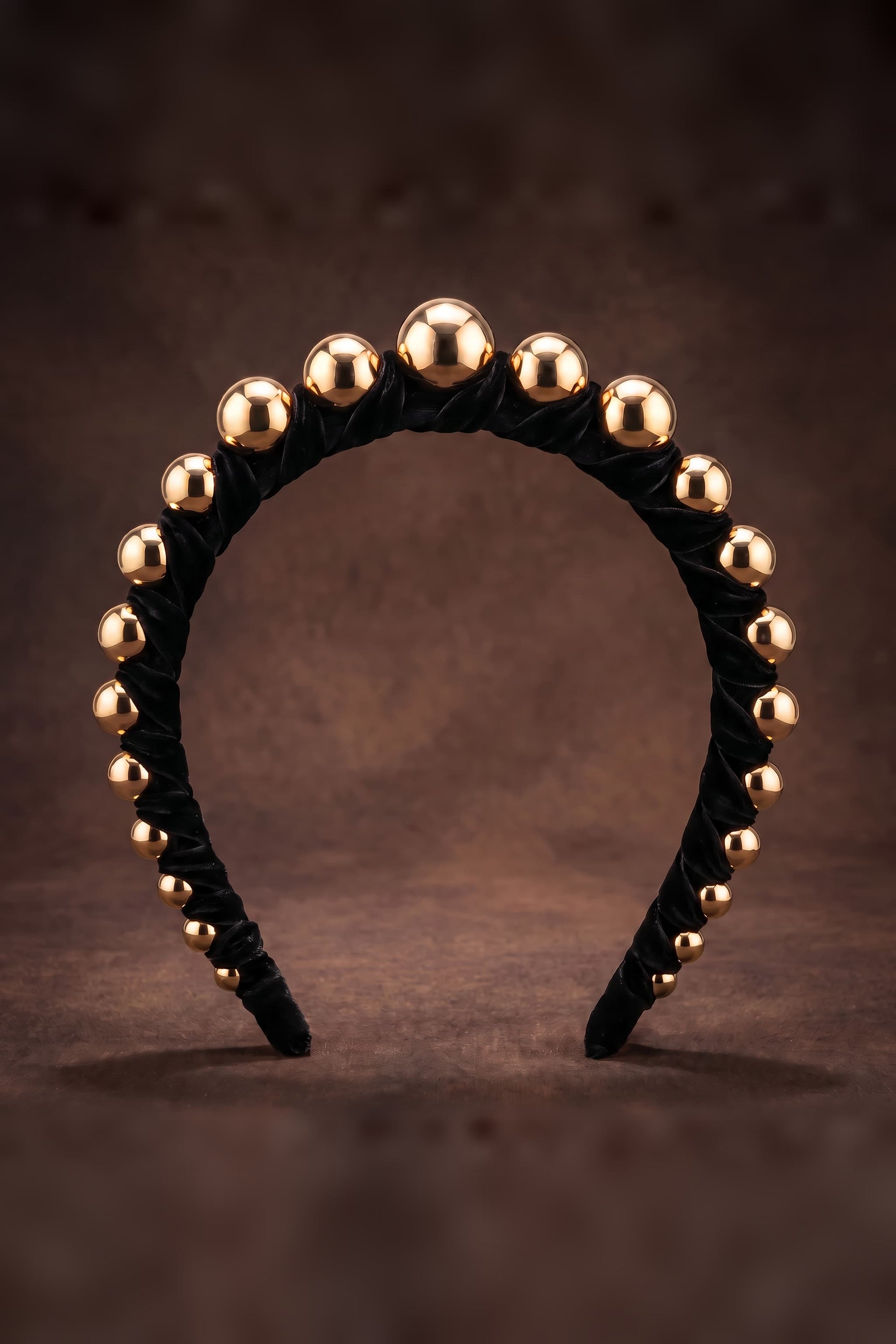 A black velvet headband decorated with a row of metallic gold spherical beads, displayed against a dark brown textured background.