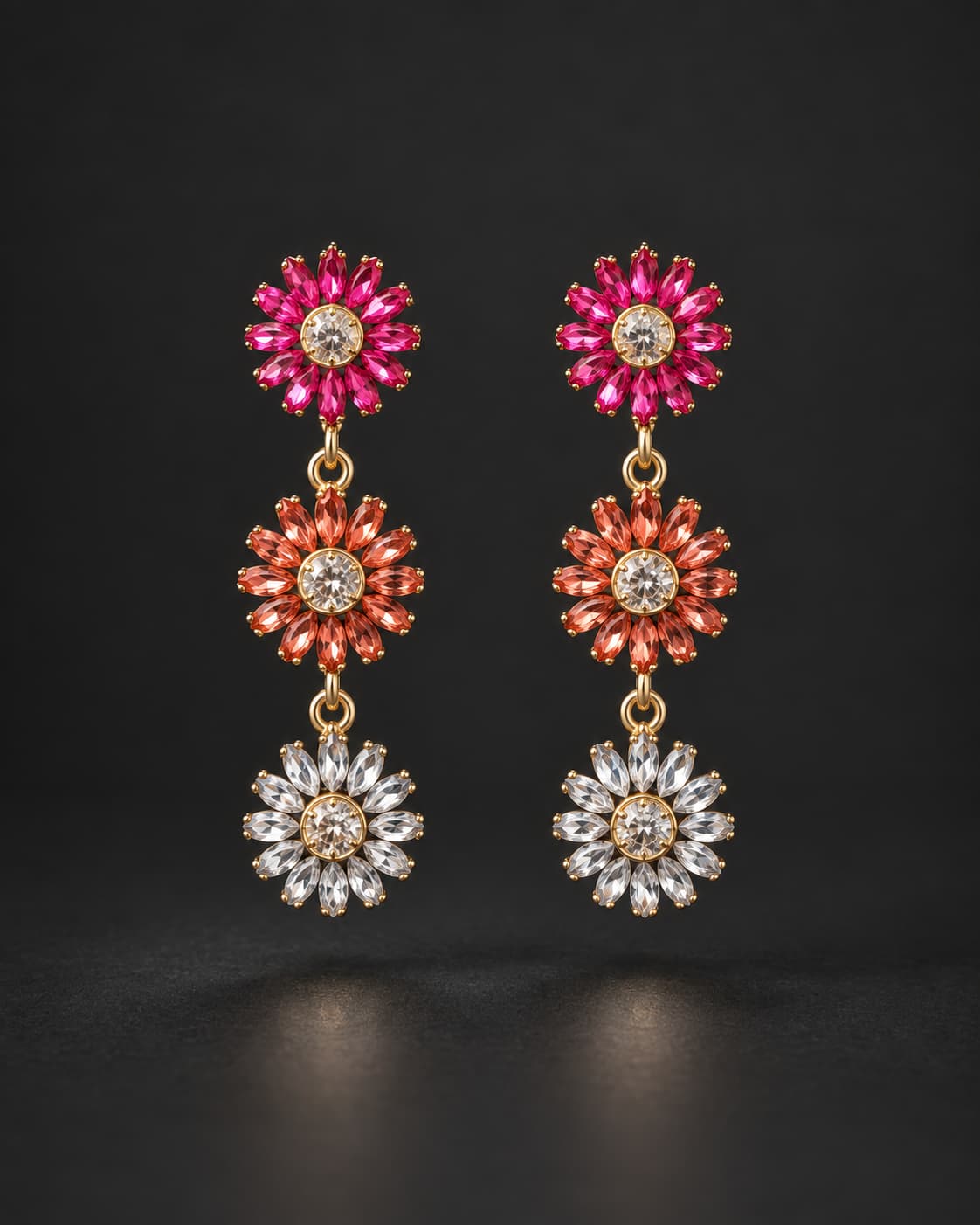 A pair of gold dangle earrings featuring three flower-shaped drops: vibrant pink, orange, and clear crystal petals, displayed against a dark background.