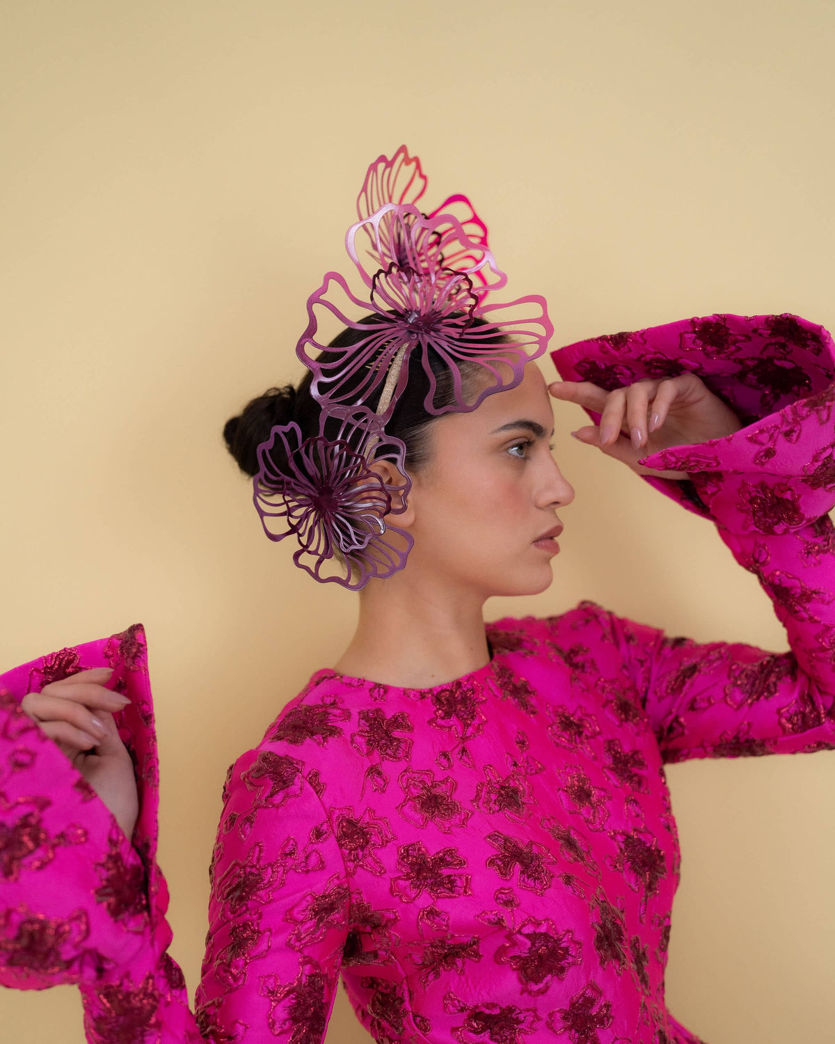 A model wearing a vibrant magenta dress with a dark floral pattern and a matching headpiece, posed against a solid beige background.