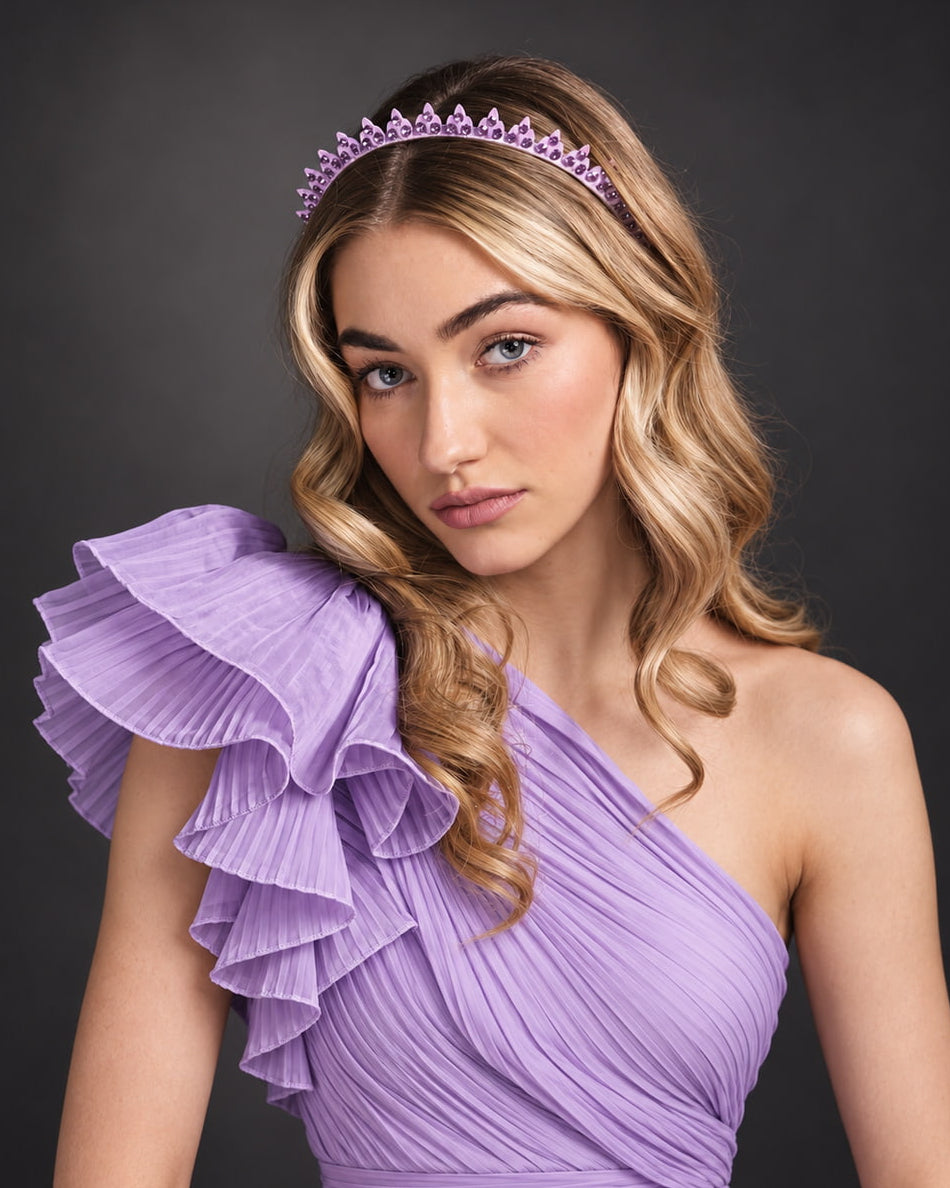 A woman wearing a lavender one-shoulder ruffled dress and a matching lavender tiara, posed against a dark gray studio background.