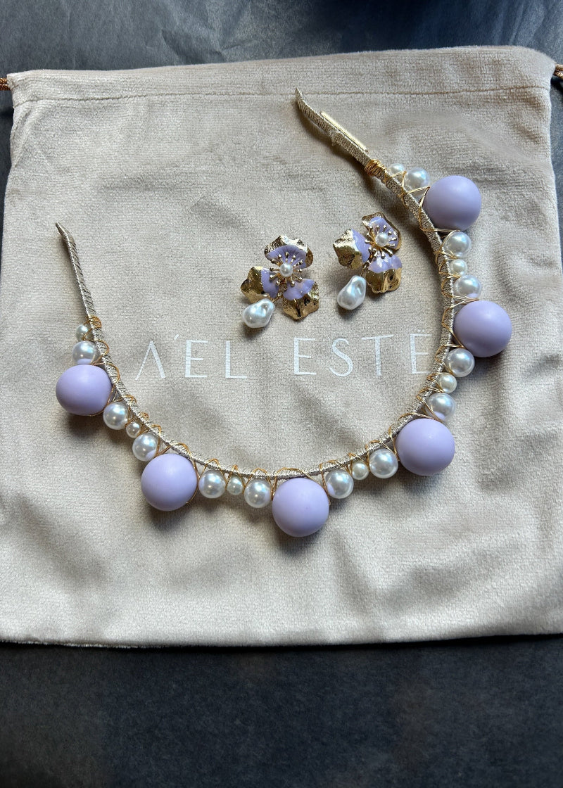 Lilac headband and earrings with light purple beads and gold accents on a 'Ael Este' branded pouch.