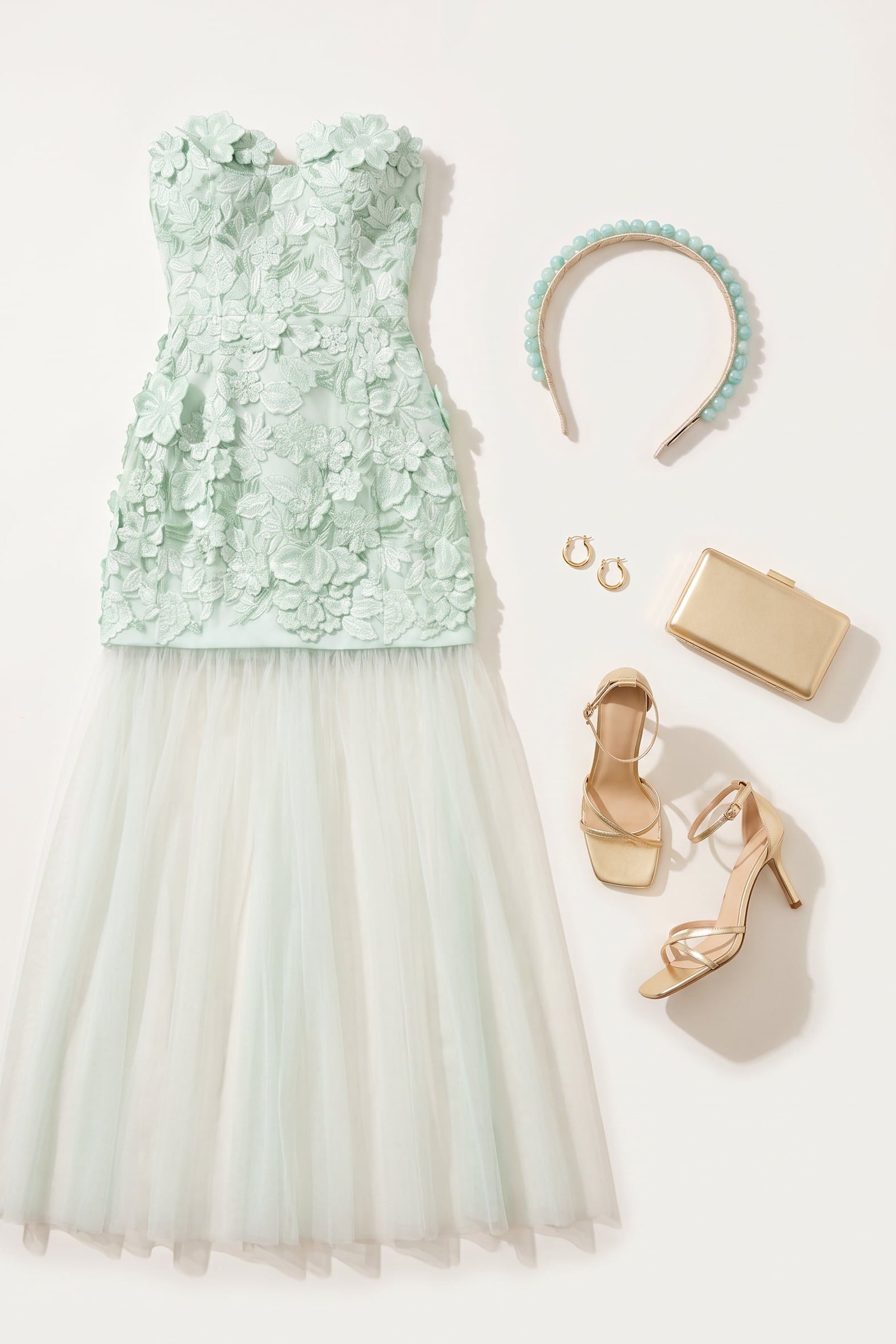 A styled flat lay of a elliatt roseate dress with a mint green floral lace bodice and a voluminous white tulle skirt, accompanied by a mint green beaded headband, gold hoop earrings, a gold clutch, and gold high-heeled sandals on a light beige background.