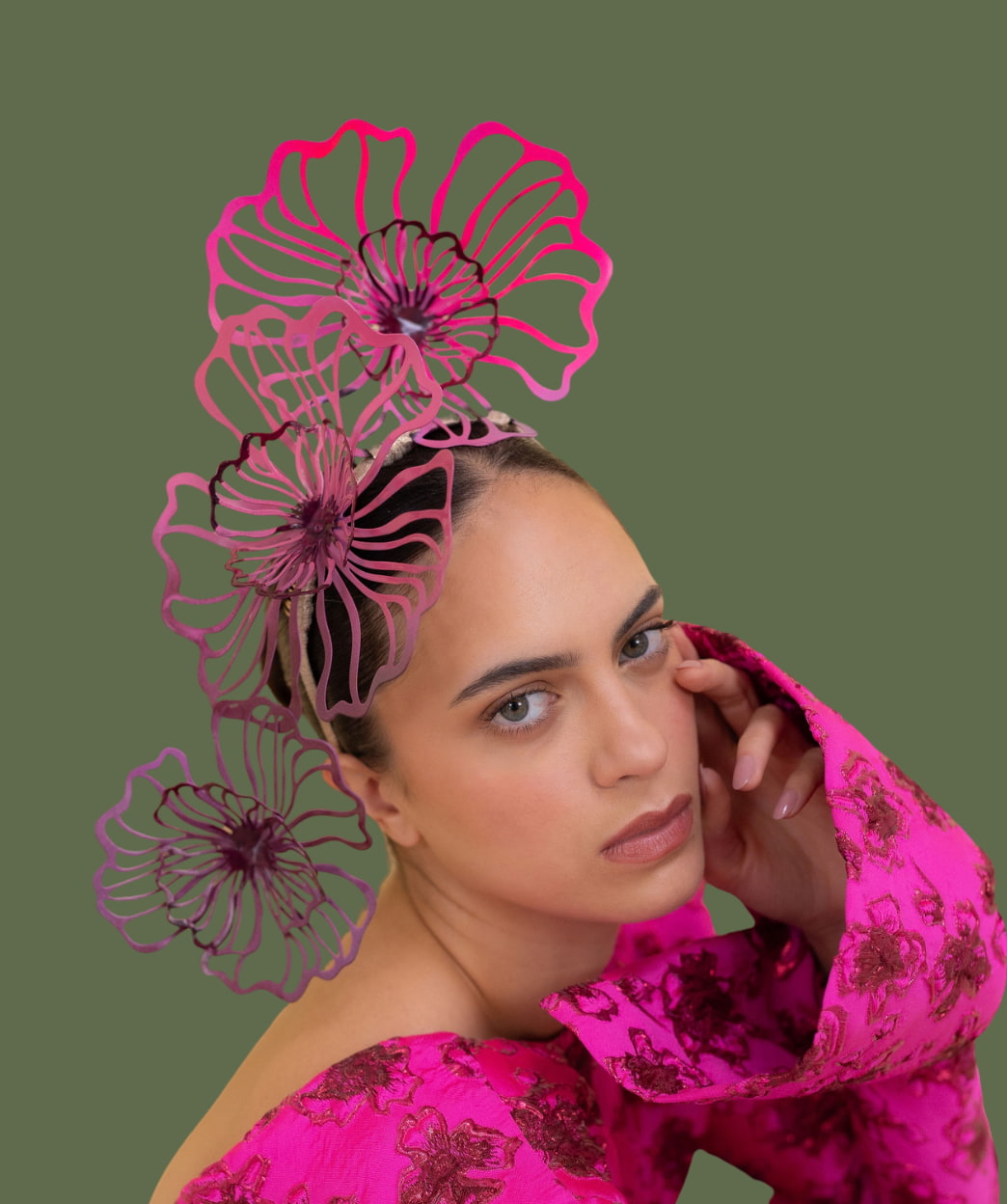 A close-up portrait of a woman wearing a dramatic hot pink floral headpiece with layered wire flower shapes and a matching bright pink patterned top, set against a solid olive green background.