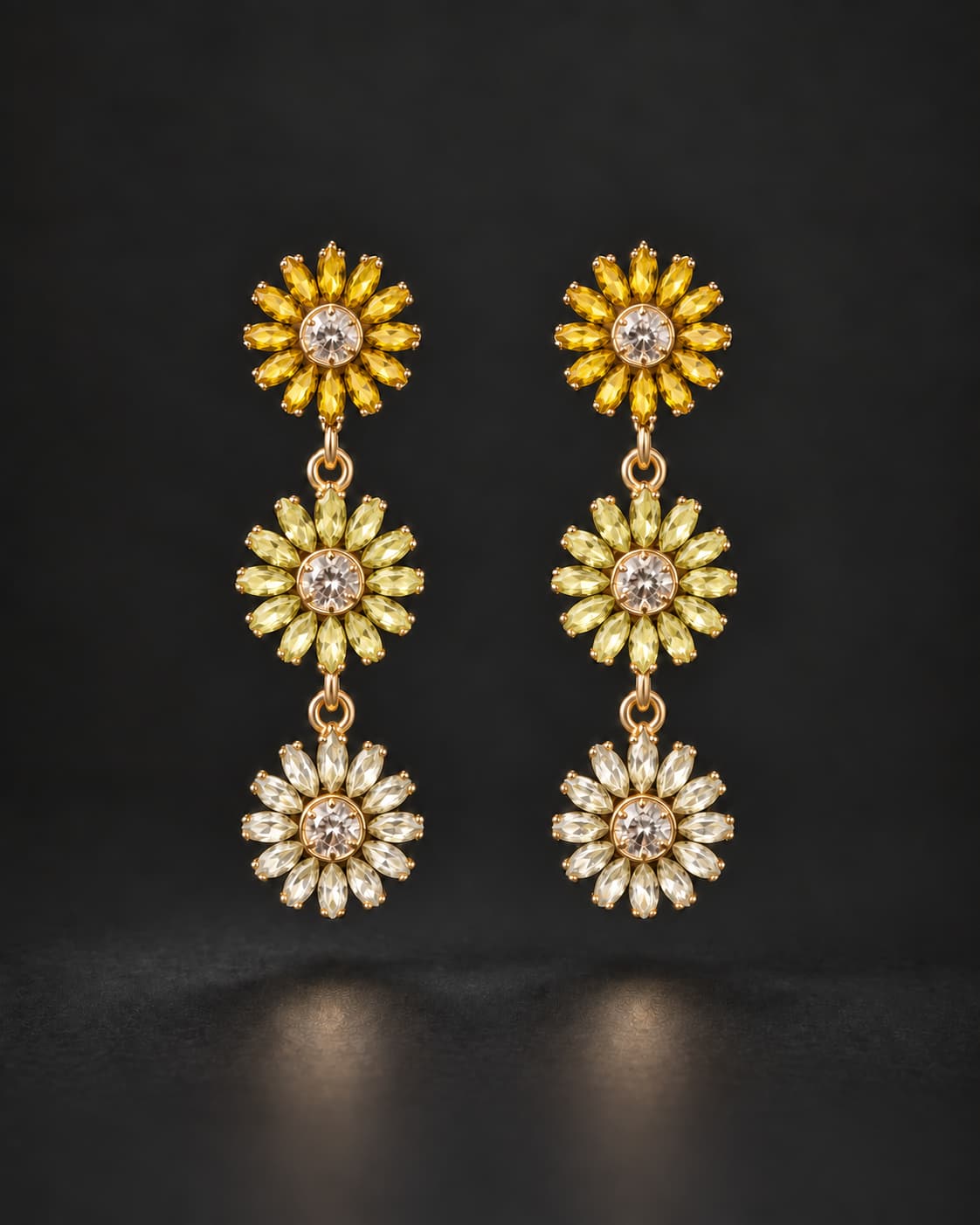 A pair of gold dangle earrings featuring three stacked sunburst flower motifs with clear gemstones, set against a dark black background.